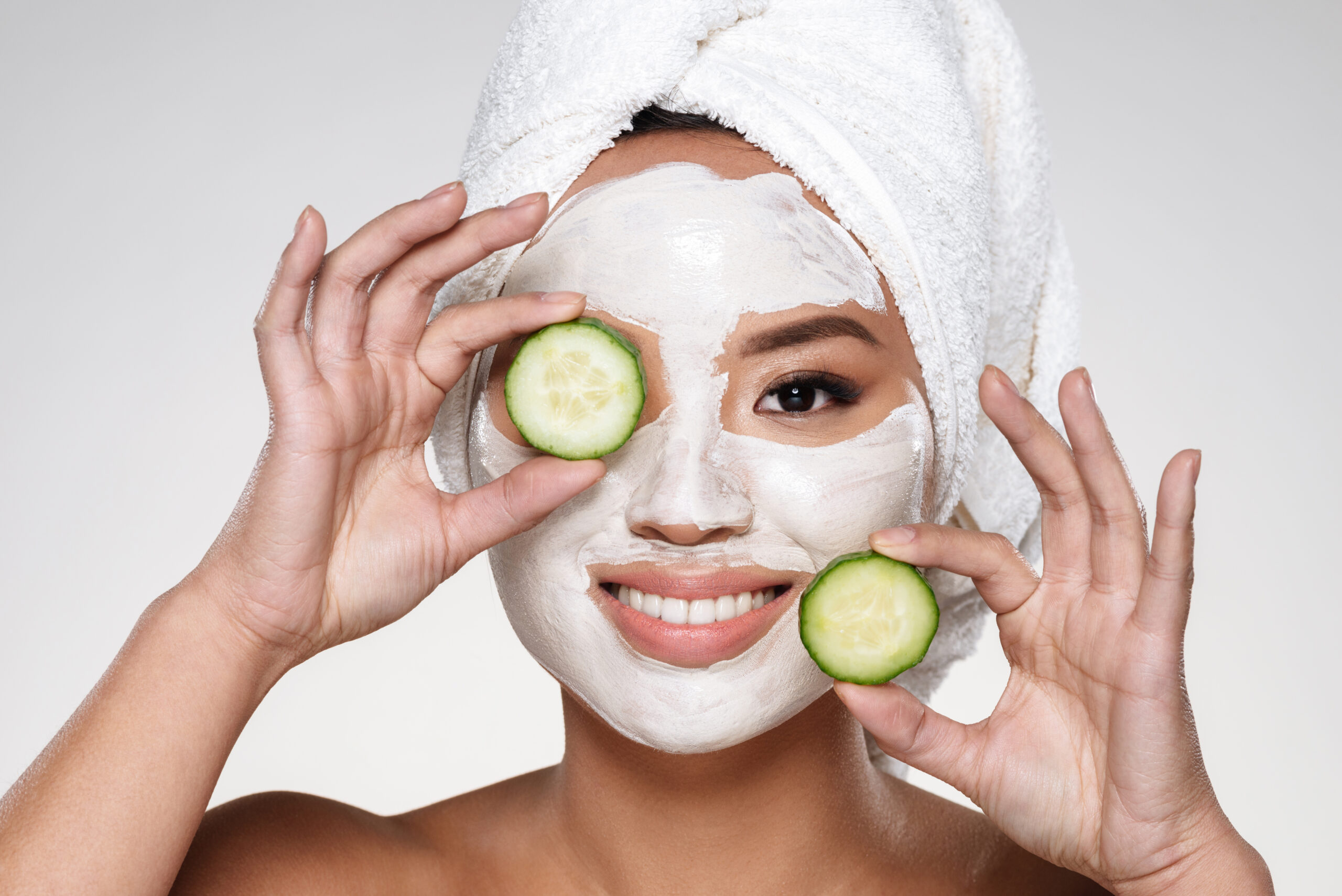 Attractive smiling asian lady with scrab on face and towel on head holding cucumber slices isolated over grey