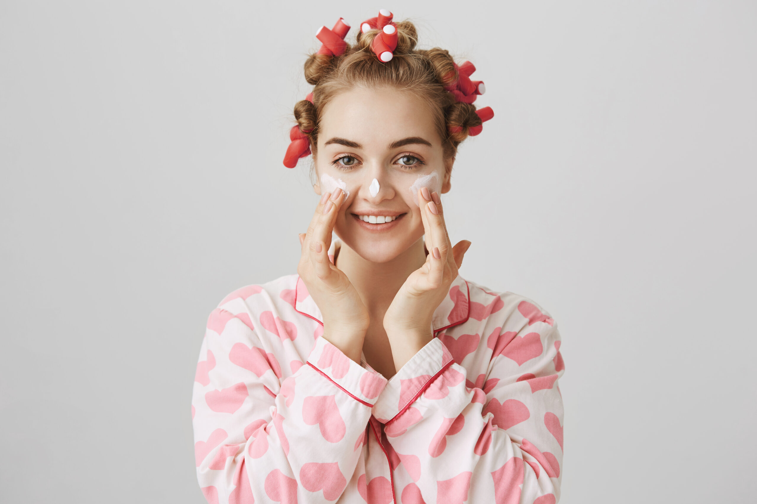 Attractive blonde caucasian model in hair-curlers, applying cream or mask on cheeks and nose, rubbing it with hands and smiling, taking care of face before going out. Skincare and beauty concept.