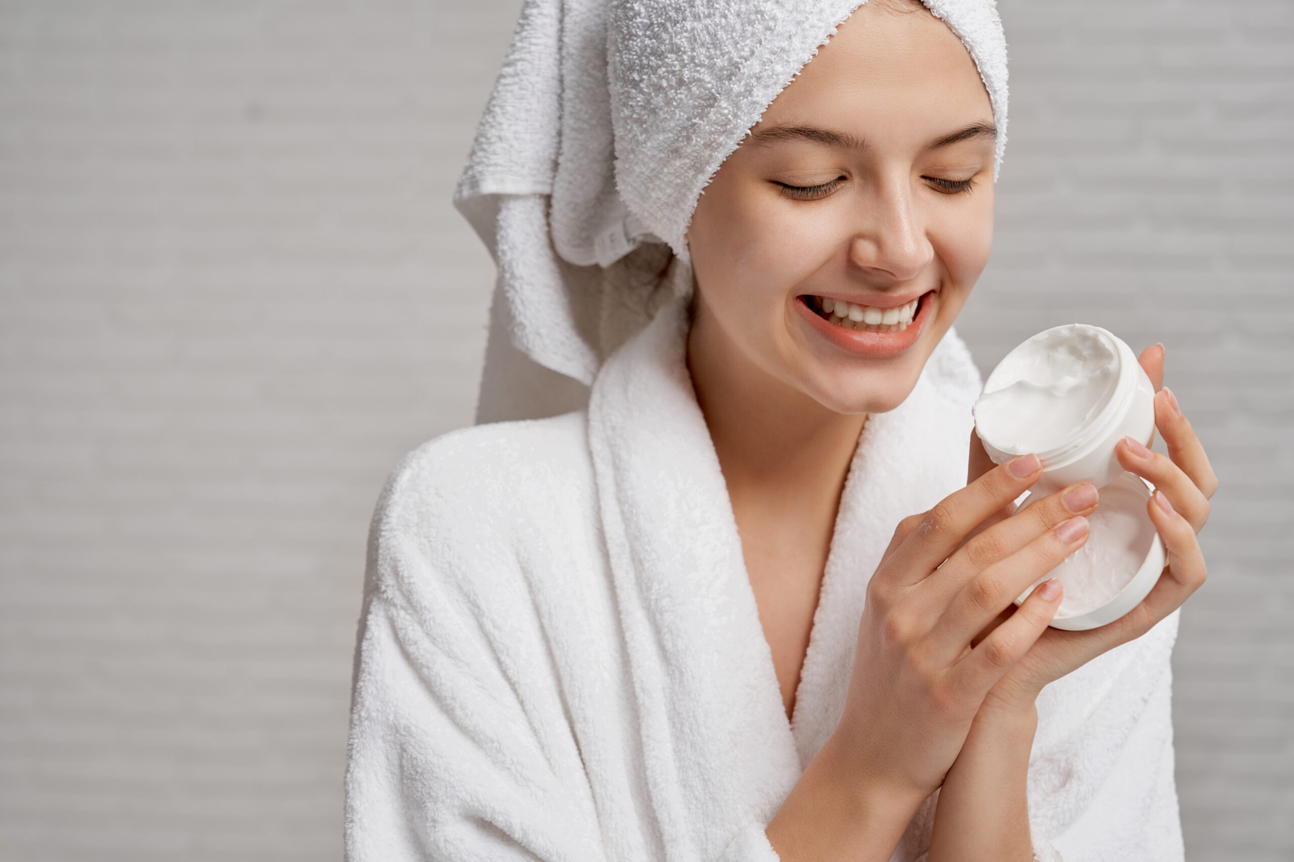 Beautiful, smiling model holding and showing box with moisturizer for face. Young woman with health, perfect skin in white bathrobe with towel on head posing on grey background.